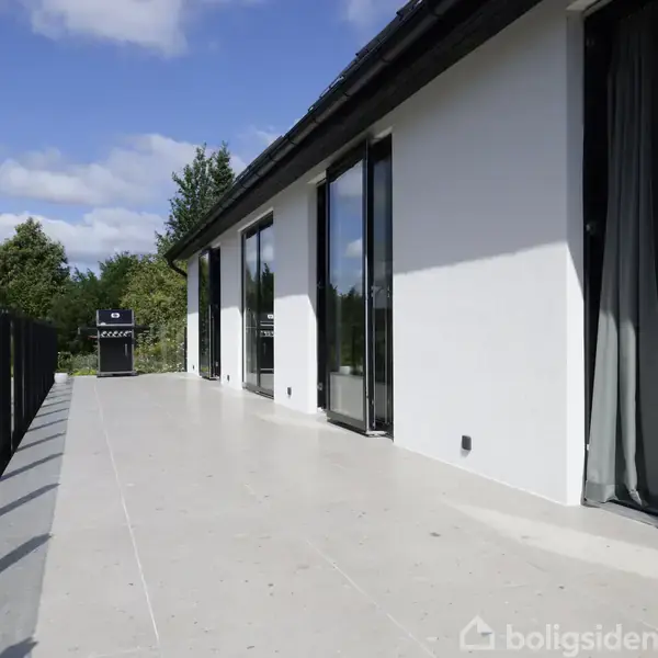 Terrassen viser en moderne bygning med store vinduer, hvor der er lagt lyse, glatte fliser. Til venstre en glasafskærmning, og i baggrunden ses grønne træer og en grill.