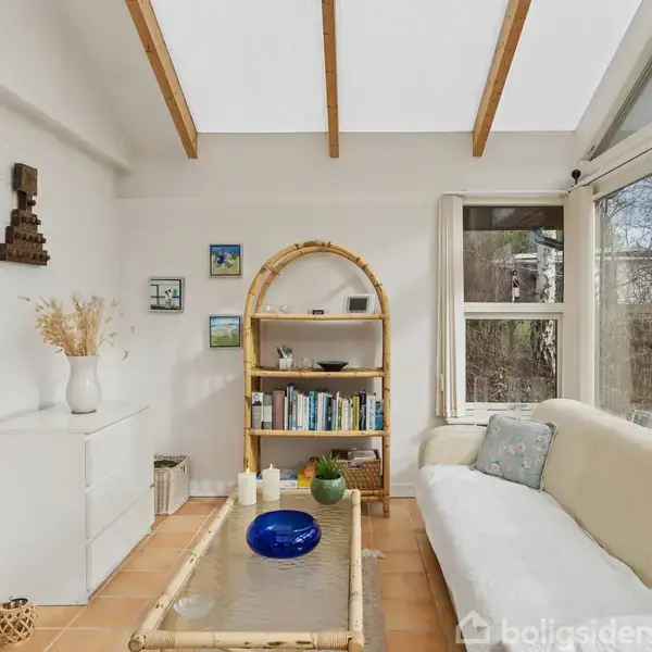 Rektangulært rum med en sofa med puder til højre, bambusbord i midten, og reol med bøger foran hvide vægge. Stort vindue til højre med udsigt til træer.