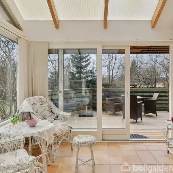 Værelse med flettede stole og bord, overdækket med blomsterstof, i en lys udestue. Store vinduer og glasdøre åbner ud mod en terrasse med siddemøbler. Fjernsyn på væggen.