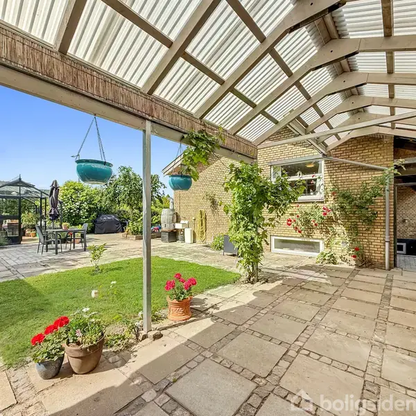 En gårdhave med fliser og græsplæne. En pergola med hængende planter dækker en del af terrassen. Et drivhus og udendørsmøbler ses i baggrunden, omkranset af frodige planter.