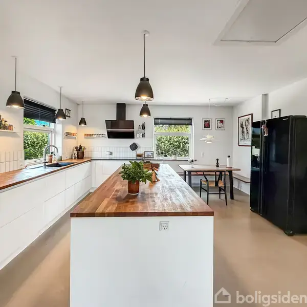 Et moderne køkken med træbordplader dominerer rummet. En plante står på køkkenøen. Sorte hængelamper hænger over bordene. Et spisebord står ved et vindue i baggrunden.