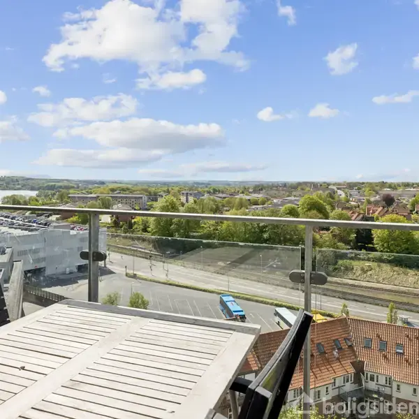 Et udendørs bord med stole står på en balkon. Udsigten viser en by med veje, bygninger og grønne områder under en blå himmel med skyer.
