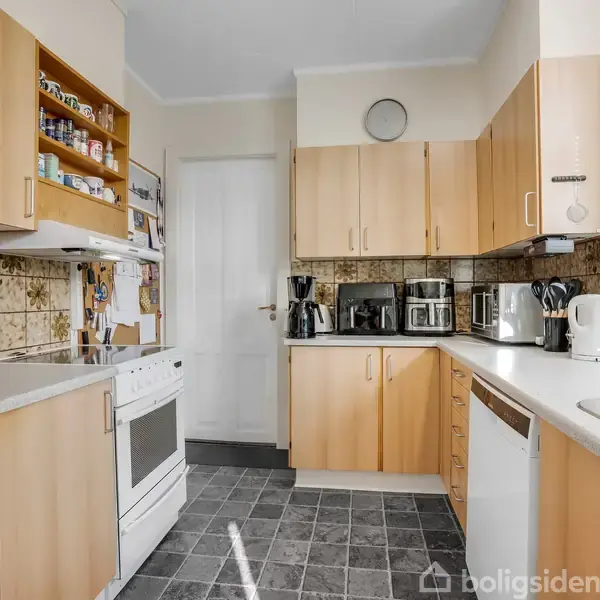 Et køkken med træskabe og hvidevarer som ovn, kaffemaskine og opvaskemaskine. På væggen er der en hylde med krydderier, og der står en plante ved vinduet.