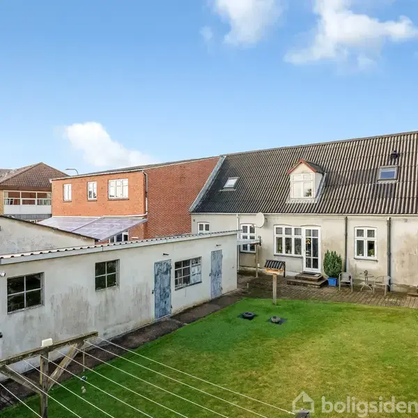 En hvid bygning med grå tagsten står stille i en indhegnet have. Området er omgivet af en betonmur og en ældre, rød murstensbygning under en blå himmel.
