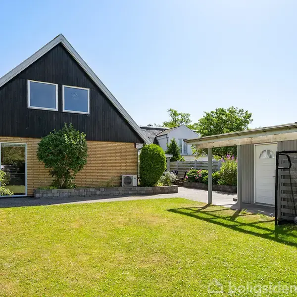 En gul murstenshus med sort tag er omgivet af en velplejet græsplæne og træer. En carport med halvtag står til højre. Sollys fylder et roligt boligområde.