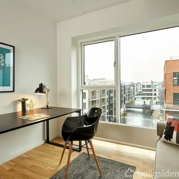 Et skrivebord med en lampe og plante står ved et stort vindue, med udsigt over en kanal og moderne bygninger. En stol er placeret ved bordet, og der hænger et billede på væggen.