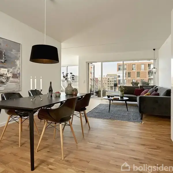 Spisebord med stole står i en moderne, lys stue. Et kunstnerisk maleri hænger på væggen. Store vinduer åbner til en altan med udsigt til byliv udenfor.