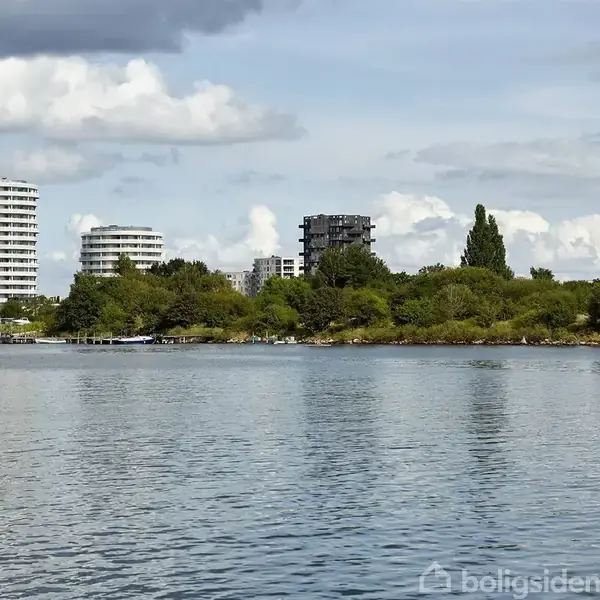 Runde moderne bygninger står stille ved en sø, omgivet af frodig bevoksning. I baggrunden hæver skyer sig i en blå, delvist overskyet himmel.