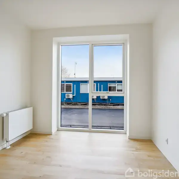 Et lyst rum med et stort vindue, der åbner ud mod en udsigt af en blå containerbygning. Et hvidt radiator står ved væggen. Gulvet er lyst træ.