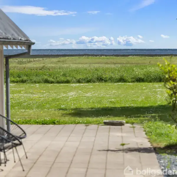 Et hus med en terrasse med stole vender ud mod en stor, grøn græsplæne, omgivet af havudsigt og blå himmel med skyer.