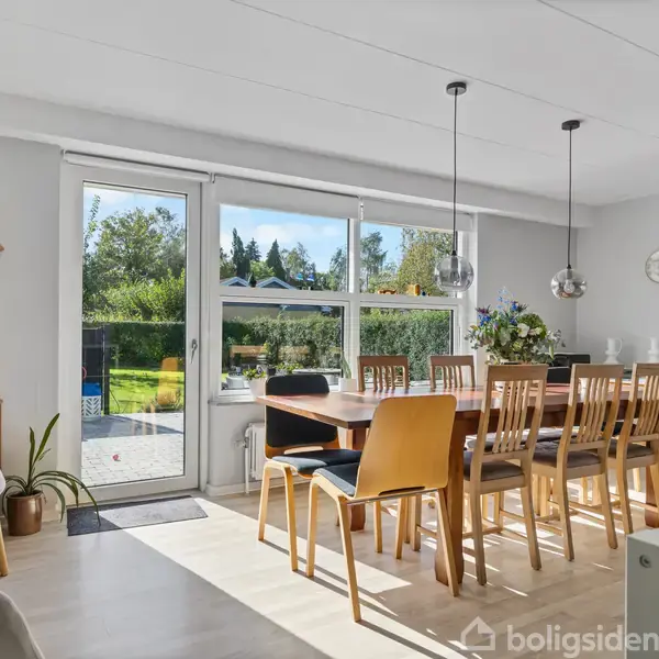 Et spisebord med træstole står i et lyst rum. Bordet er dækket med en vase med blomster. Store vinduer og en dør fører ud til en grøn have. En trækommode og dekorative elementer er i rummet.