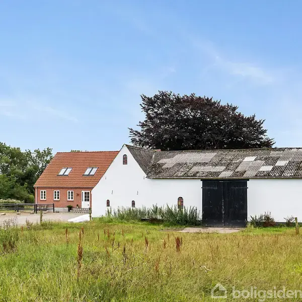 En hvid lade med sort dør står foran en rød murstensbygning med skråt tag. Græsarealer og træer omgiver bygningerne under en blå himmel.