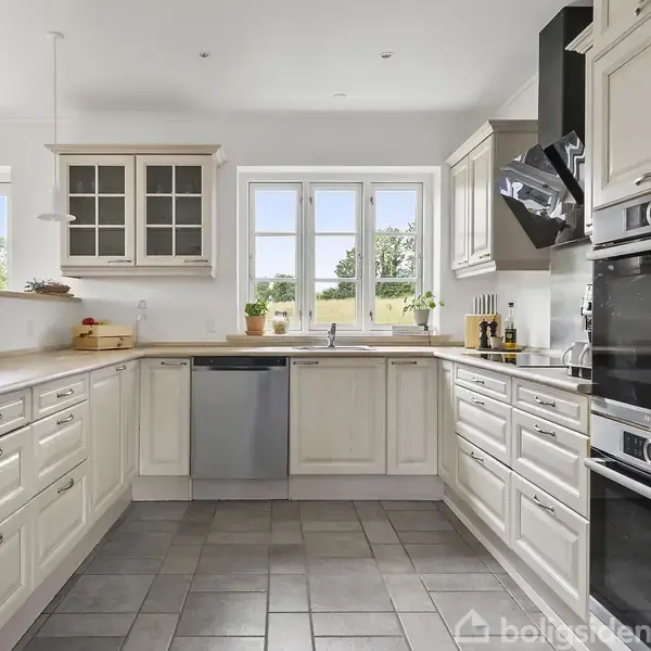 Køkken med hvide skabe, opvaskemaskine og dobbeltovn; stort lysindfald fra vinduer ud til grøn udsigt; moderne indretning med lyst træbordplade og grå flisegulv.