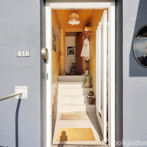 Dør står åben, fører ind til en lys gang med pastelgule vægge og planter. På væggen står: "Hjemmelavede kager rugager pålæg". Husnummer 333 vises til venstre for døren.