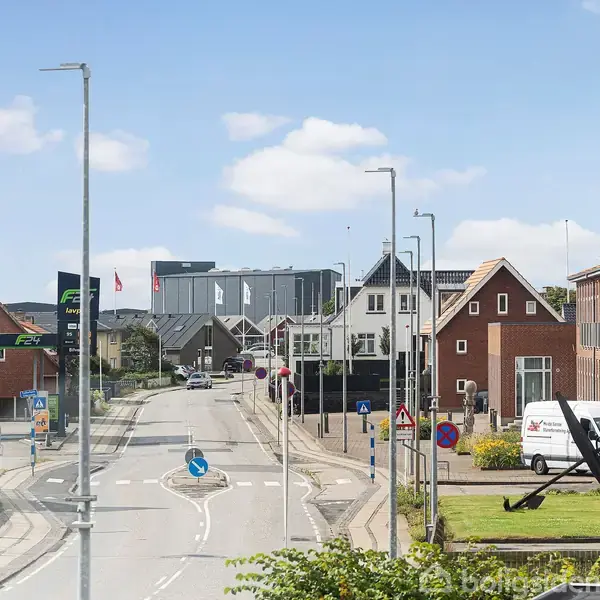 En stille gade med små huse og en varevogn parkeret langs vejen. Et kunstværk er placeret på græsset. Skyfri himmel skaber en klar og solrig dag.