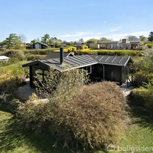 Et sort træhus står omgivet af tæt buskads og græsplæne. I baggrunden ses flere huse, træer og buske under en klar blå himmel.