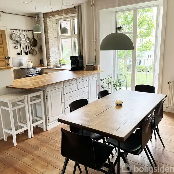 Køkkenalrum med lang træbord omgivet af sorte stole. Hvid køkkenø med barstole står ved siden af. Store vinduer viser en grøn udsigt. Væghængte køkkenredskaber og planter dekorere rummet.