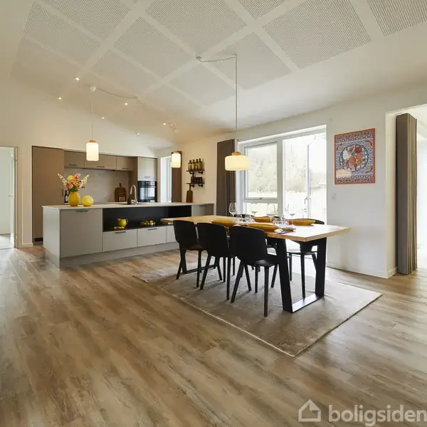 Stort, lyst køkken med moderne indretning, hvor et spisebord med sorte stole står under en hængende lampe. Køkkenøen har blomster og frugter. Store vinduer giver udsigt til det fri.