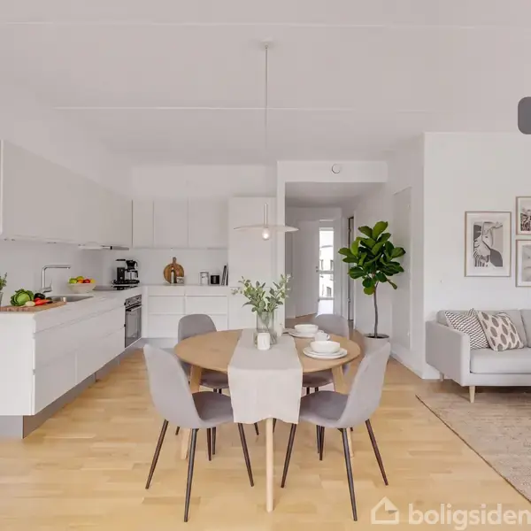 Et lyst køkkenalrum med et spisebord og fire stole i midten, omgivet af en moderne køkkensektion til venstre og en sofa med billeder på væggen til højre. Text: "Visualisering".