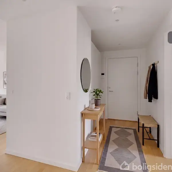 En lys entré med et rundt spejl og en træbænk ved siden af en dør. Et tæppe ligger på gulvet, og der er indrettet med planter og jakker på knager. I baggrunden ses en stue med sofa og sofabord.