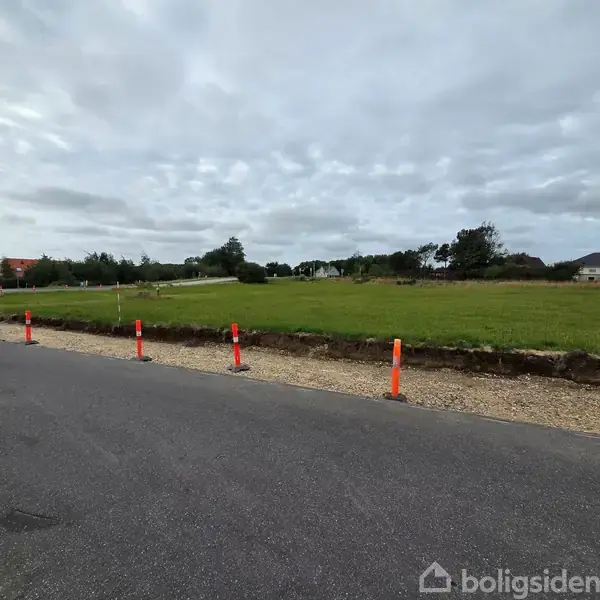 En række orange kegler står langs en grusstribe ved vejkanten. Området består af en åben grønt græsmark, omkranset af spredte træer under en overskyet himmel.