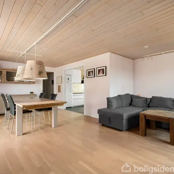 Stue med grå hjørnesofa og træbord til højre, bord og stole til venstre under to lamper. Lys trægulv og træloft. Væghængt reol og køkken i baggrunden.