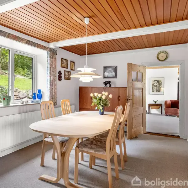 Træbord omgivet af fire stole står i en spisestue med træloft. Bordet har en vase med blomster. Vinduet viser en grøn have. En åben dør fører til et andet rum.