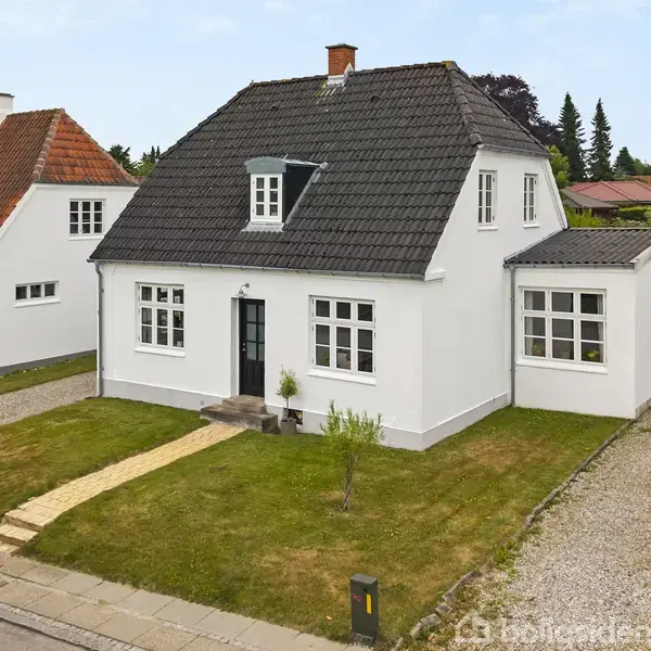 En hvid murstensvilla med sort tag ligger ved en villavej. Den har en sti, græsplæne og grusindkørsel med garage. Naboer ses til venstre.