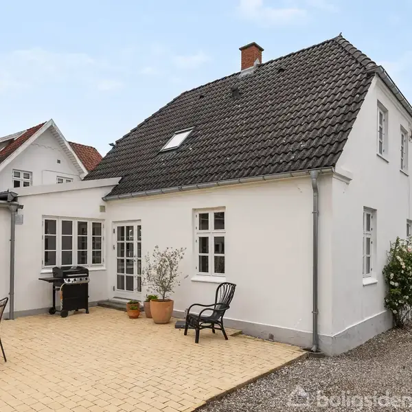 Hvidt hus med mørkt tag står i et gårdmiljø med flisebelægning. Havemøbler og en grill er placeret rundt om gårdspladsen, med grønne planter i nærheden.