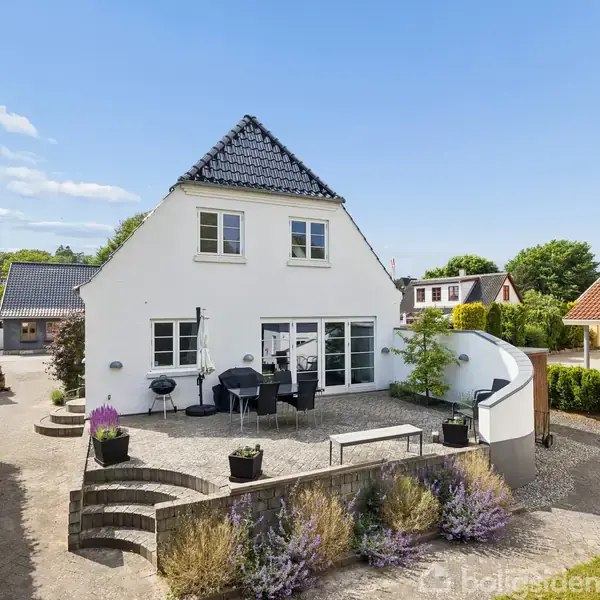 Huset har hvid facade og sort tag, omgivet af en solrig terrasse med møbler og planter. Det ligger i et kvarter med lignende huse, omgivet af grønne buske og træer.