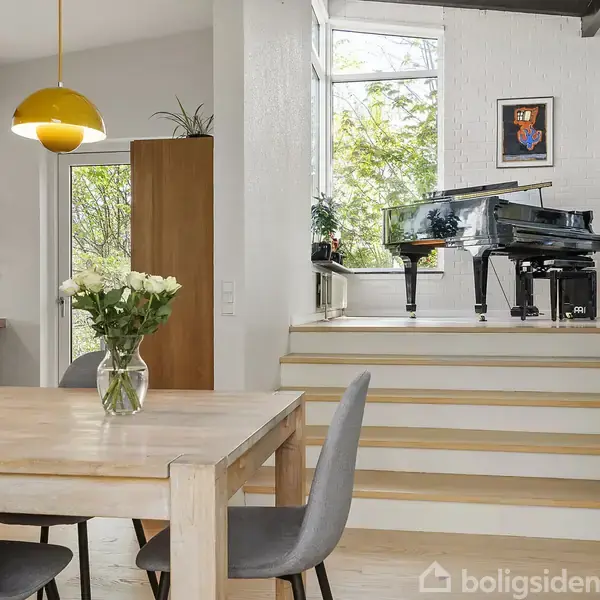 Et spisebord med blomster på står i køkkenalrum. Ovenfor trappen står et sort flygel i et lyst rum med store vinduer og kunst på væggene.