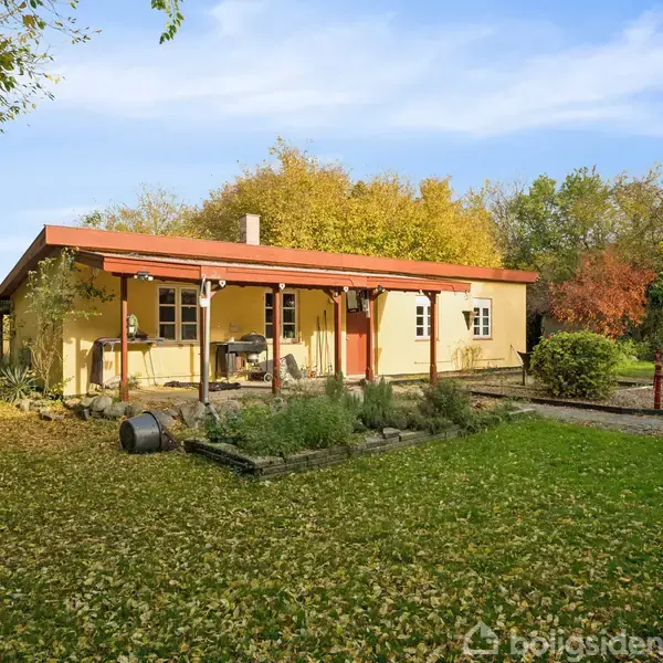 Et lille gult hus med rødt tag står omgivet af en frodig have med træer og græsplæne. Huset har en veranda med stole og bord.