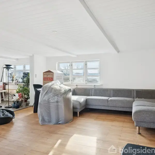 En stor grå hjørnesofa står i en rummelig stue med trægulv. En moderne brændeovn er placeret i hjørnet. Vinduerne giver naturligt lys. Møbler og flyttekasser er til stede.