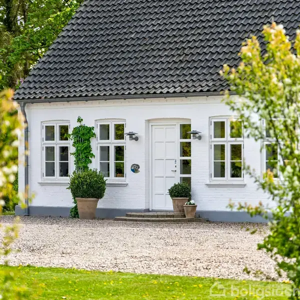 Hvidmalet hus med sort tegltag står blandt grønne buske og træer. To store potteplanter ved indgangspartiet. Husets dør er hvid med flere vinduer omkring. Gruset indkørsel foran.