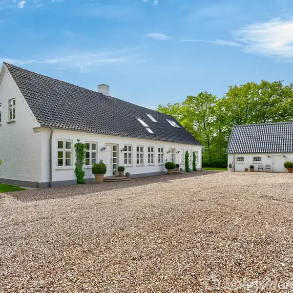 Et hvidt landhus med sort tag står stille i en grusbelagt gård, flankeret af en mindre bygning. Omgivet af grønne træer under en blå himmel.