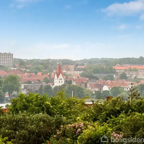 Kirke med rødt tag står midt i en by med mange røde bygninger, omgivet af grønne træer og buske, under en klar blå himmel.