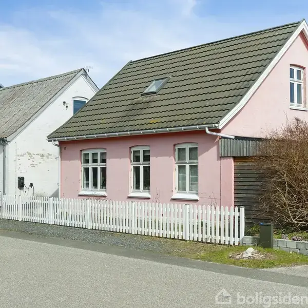 Et lyserødt hus med hvide vinduer og et gråt tag står ved siden af et hvidt hus. Huset har en hvid stakit og busk i haven.