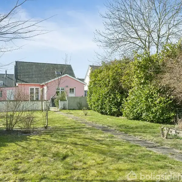 Et lyserødt hus med sorte tagsten står i baggrunden. En grøn græsplæne omgivet af buske og træer danner haven, med en stensti der fører mod huset.