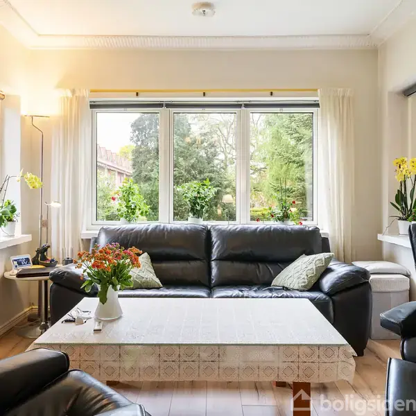 Sofa i sort læder med puder står foran et stort vindue med udsigt til en have. Et sofabord med blomster står i midten af et lyst, indbydende rum.