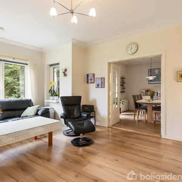 En stue med en sort sofa og lænestol omkring et bord med blomster. Store vinduer giver lys, mens en åben dør fører til et spisestueområde. En væg har en ur.