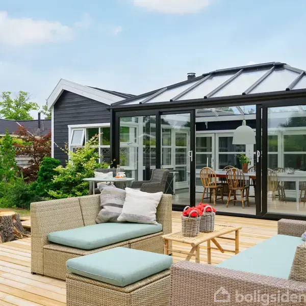 En udendørs terrasse med sofamøbler og puder står ved siden af et glashus, hvor der er et spisebord med stole. Omgivet af træer og buske i en haveindretning.