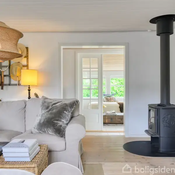 En stue med sofa og puder står foran en brændeovn. Et bord med magasiner og planter er ved siden af. Rummet har lyse vægge og gulv, med dør til et andet rum.