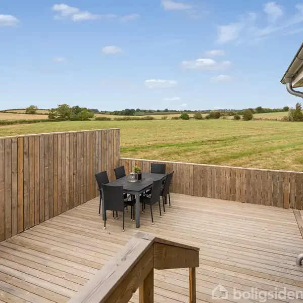 Terrasse med træmøbler står på et stort trædæk, omgivet af en lav trævæg og grønne marker i baggrunden under en blå himmel.