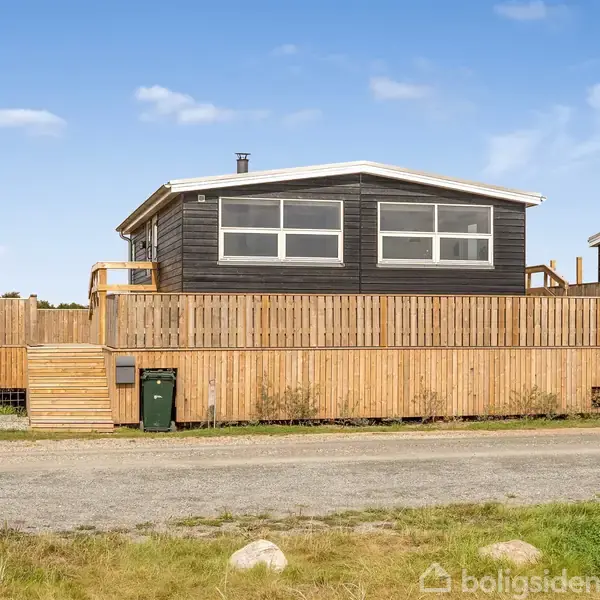 Et sort træhus står stille bag et højt trægærde. I forgrunden ses en grusvej og en grøn skraldespand. Huset er omkranset af en klar blå himmel.