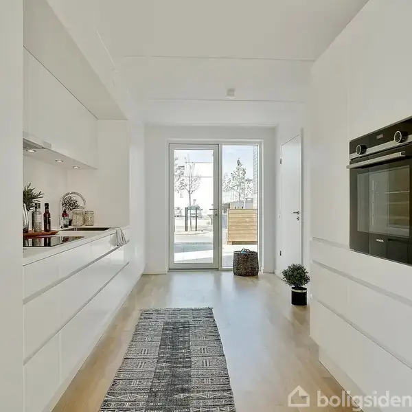 Et moderne, lyst køkken med indbygget ovn til højre. Et tæppe ligger på gulvet, og en glasdør fører ud til en gårdhave. Pynt omfatter planter og køkkenudstyr.