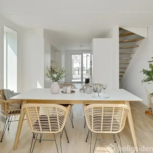 Et minimalistisk spisestue med en hvid bordplade og fire fletstole. En vase med planter dekorerer bordet. Til højre er en trappe, og omgivelserne er lyse og moderne.