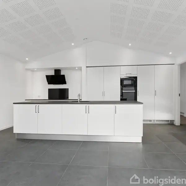 Hvidt køkken med moderne køkkenø, udstyret med indbygget kogeplade. Væggene er hvide, gulvet gråt, omgivet af skabsvægge og integrerede apparater i minimalistisk stil.
