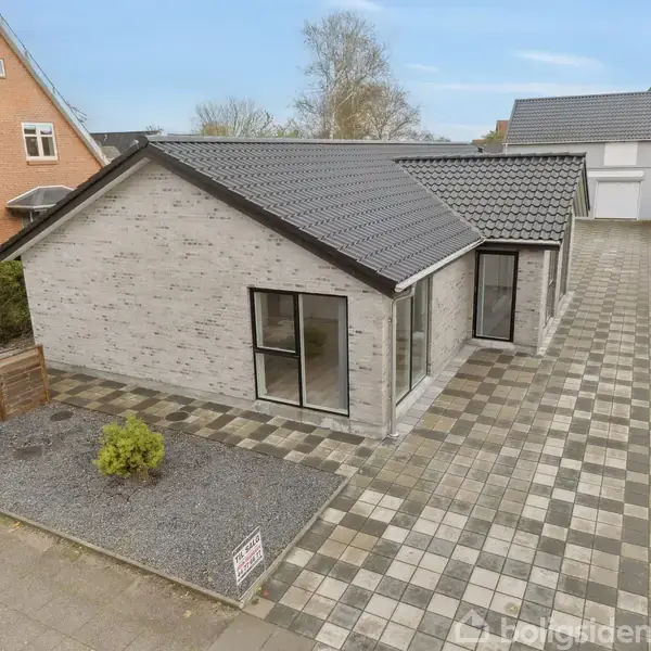 En etplans stuehus med gråt tegltag står overfor et flisebelagt område og en lille have. Husets facade er grå mursten. En grøn busk er synlig ved siden af huset.