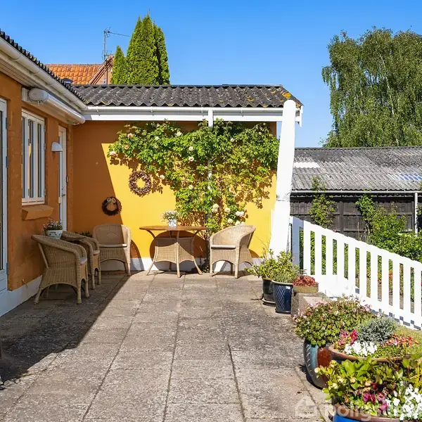 En terrasse omgivet af en gul mur. To kurvestole og et bord står under en frodig plante. Krukker med blomster pynter området. Hvide rækværk omkranser siden mod haven.