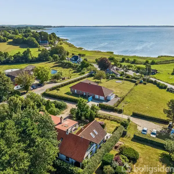 Huse ligger tæt ved kysten, omgivende af grønne marker og træer. Havet strækker sig ud i horisonten, mens solen skinner på det blå vand. Ingen tekst fundet.
